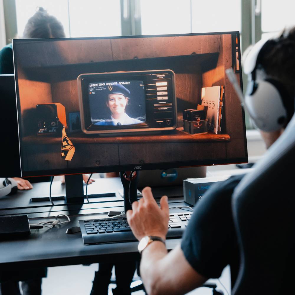 Gaming-Screen Ein Mann sitzt vor einem Computer und spielt ein Spielt. Er ist von hinten und leicht seitlich zu sehen.Vor allem ist der Bildschirm zu erkennen. Darauf ist im Spiel ein Bildschirm mit einer Frau in Uniform zu sehen.