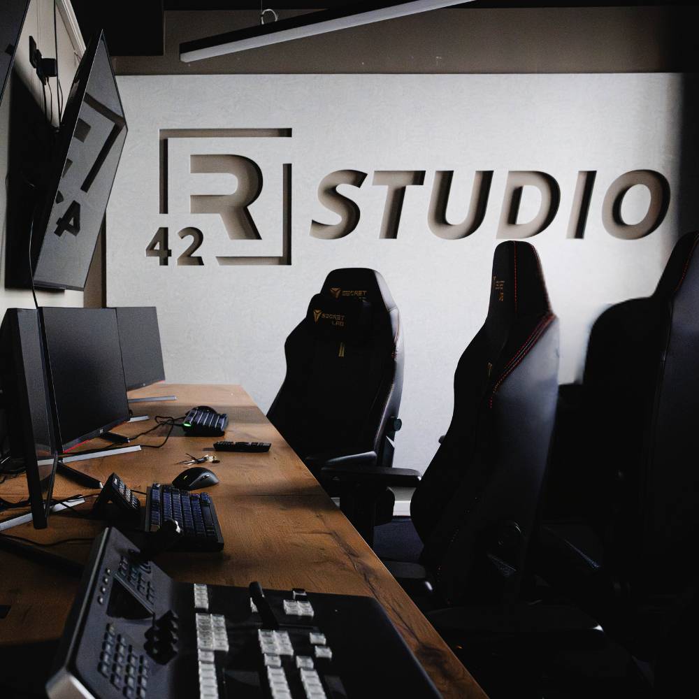 R42 Studio mit einer grauen Wand in der das Logo "R42 Studio" gestanzt ist. Im Vordergrund stehen mehrere Stühle und Aufnahmetechnik.