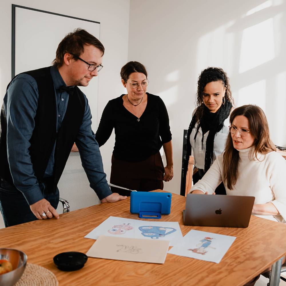 4 Personen tauschen sich in einem Workshop aus. 1 Person sitzt an einem MacBook, die 3 anderen stehen daneben und schauen auf den Bildschirm.