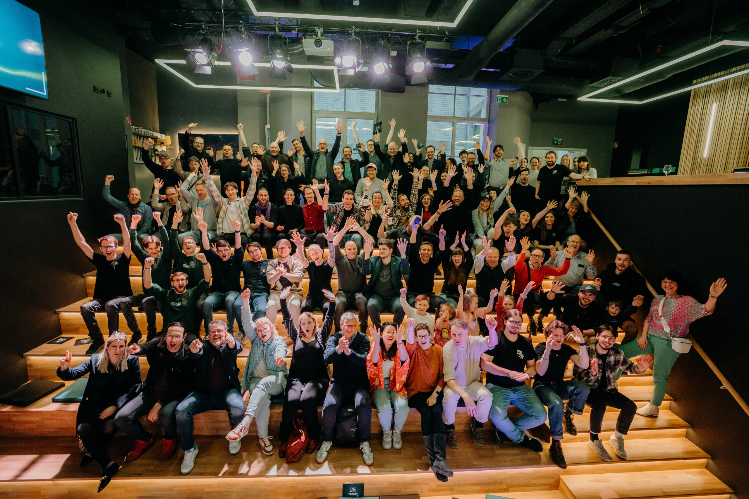 Die Teilnehmerinnen und Teilnehmer der neuen Start-up-Klasse 2025 sowie das Coaching-Team und Partnerinnen und Partner sitzen auf den Stufen der Event-Arena im R42 und heben jubelnd die Arme hoch.