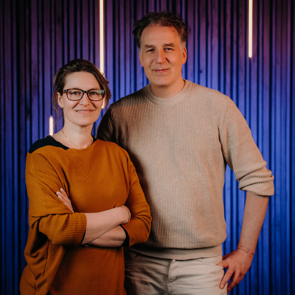Start-up – Feynarock_1000x1000 Eine Frau in einem braunen Pullover und ein Mann in einem beigefarbenen Pullover stehen nah beieinander mit Blick Richtung Kamera. Die Frau steht leicht schräg vor dem Mann und ihre Arme vor dem Bauch verschränkt.