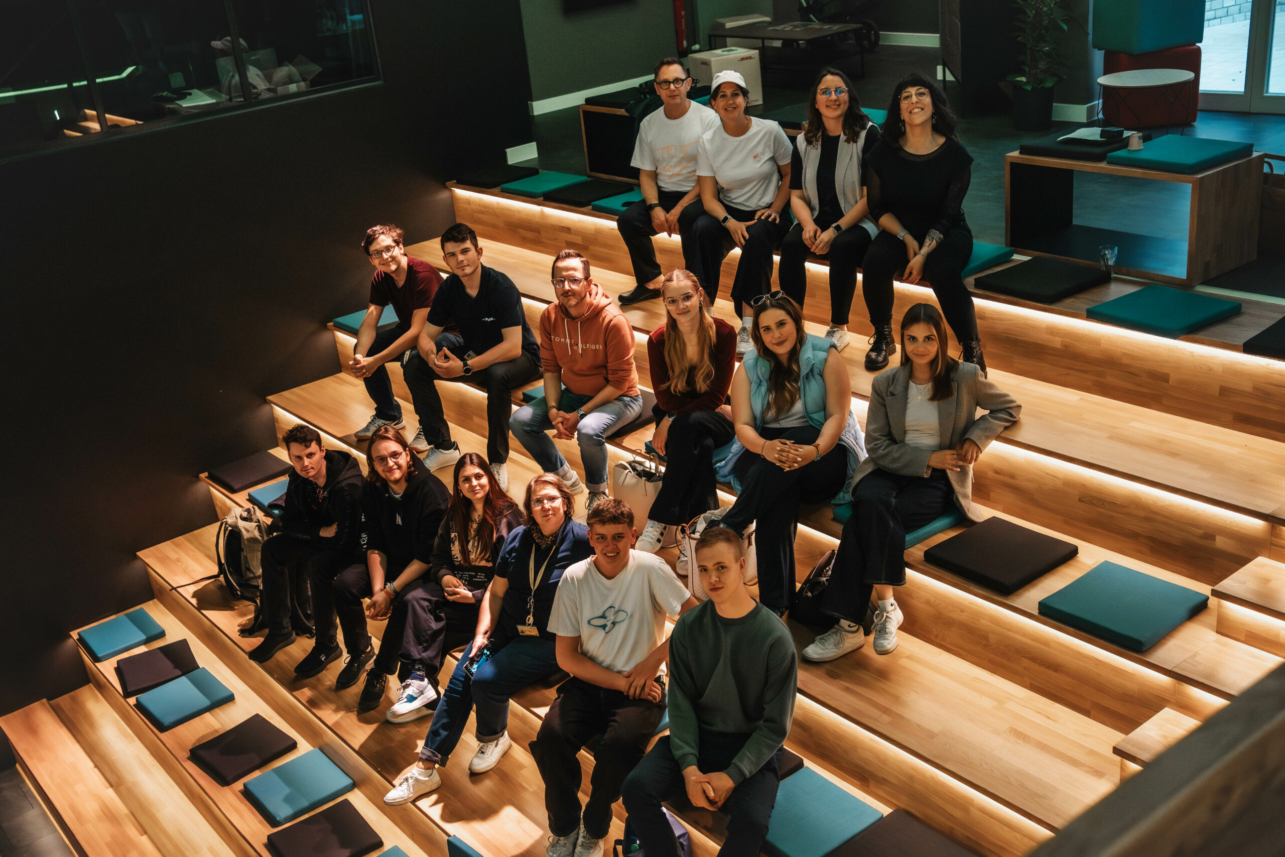 Zwölf Auszubildende und Studierende von DHL sitzen mit den Gründern von Trustbox und zwei Mitarbeiterinnen vom R42 Games Accelerator in der Eventarena des R42. Alle gucken freundlich in die Kamera. Sie sind von leicht oben fotografiert.