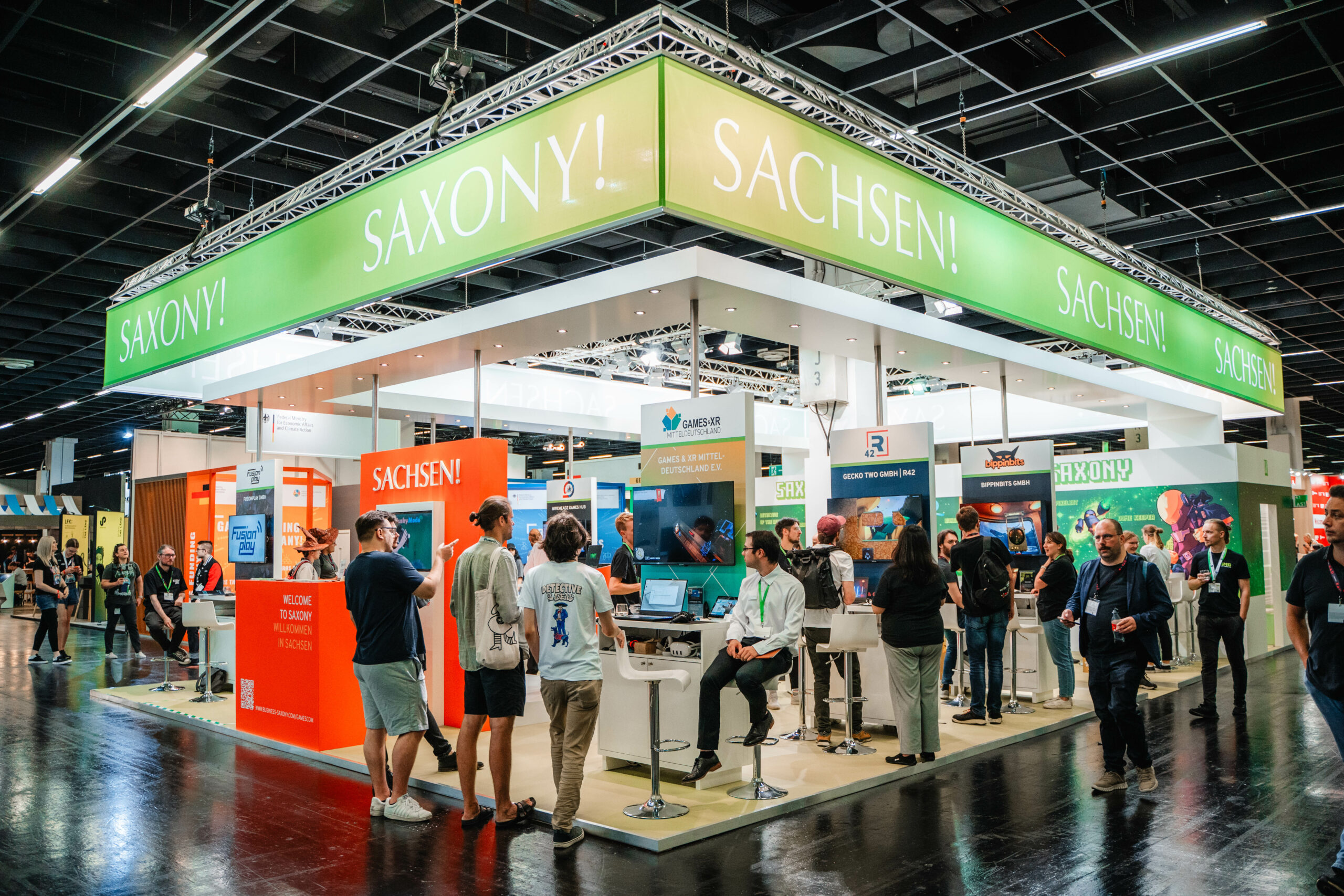 Blick auf den Messestand des Freistaates Sachsen auf der gamescom 2024. Mehrere Personen stehen vor und auf dem Stand. SIe unterhalten sich oder betrachten das Angebot der Ausstellenden.