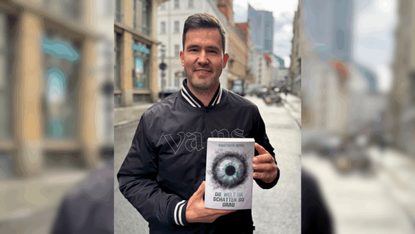 Ein braunhaariger weißer Mann Mitte 30 trägt eine schwarze Jacke mit der Aufschrift Vans. Er lächelt. In den Händen hält er das Buch "Die Welt im Schatten so grau" von Constantin Dupien und präsentiert es der Kamera. Er steht auf einer Straße in der Leipziger Innenstadt. Im Hintergrund ist das Unihochhaus zu sehen.