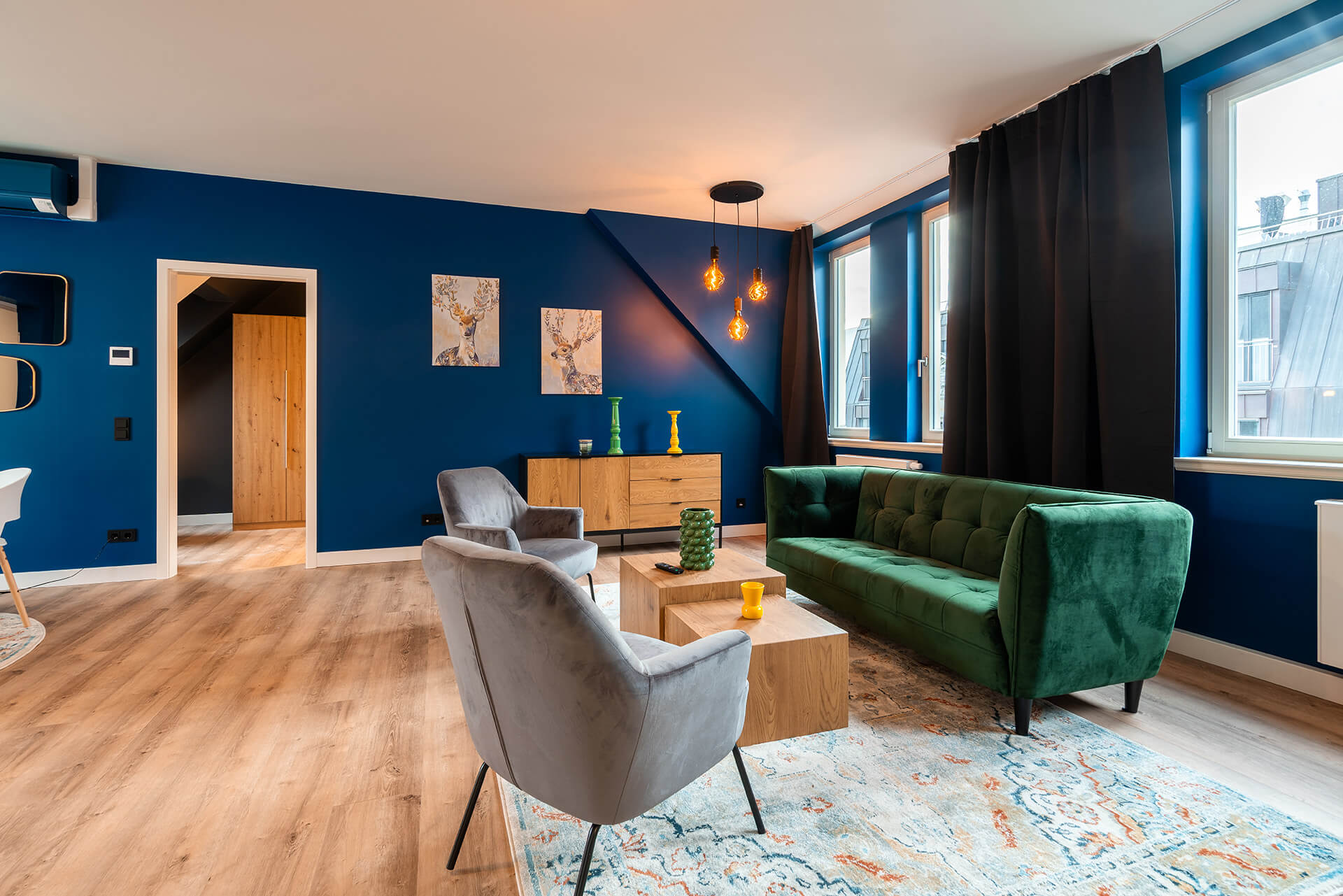 Modern eingerichtetes Apartment namens Lara's Loft im R42 in Leipzig, mit offenem Wohnbereich, grünem Samtsofa, zwei grauen Sesseln, großem Teppich, hellem Holzfußboden sowie angrenzendem Schlafzimmer.