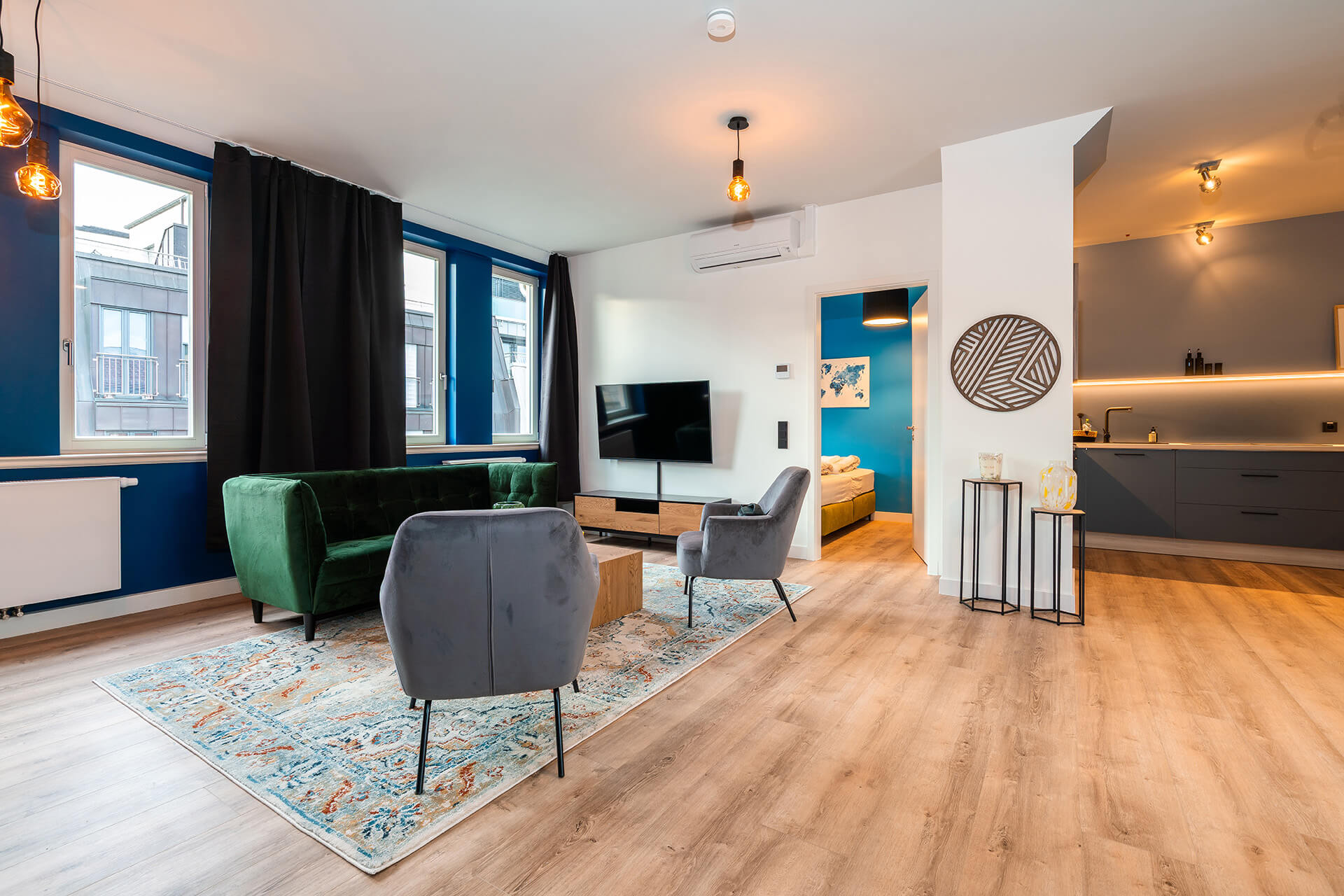 Modern eingerichtetes Apartment namens Lara's Loft im R42 in Leipzig, mit offenem Wohnbereich, grünem Samtsofa, zwei grauen Sesseln, großem Teppich, Wand-TV, hellem Holzfußboden sowie angrenzender Küche und Schlafzimmer.