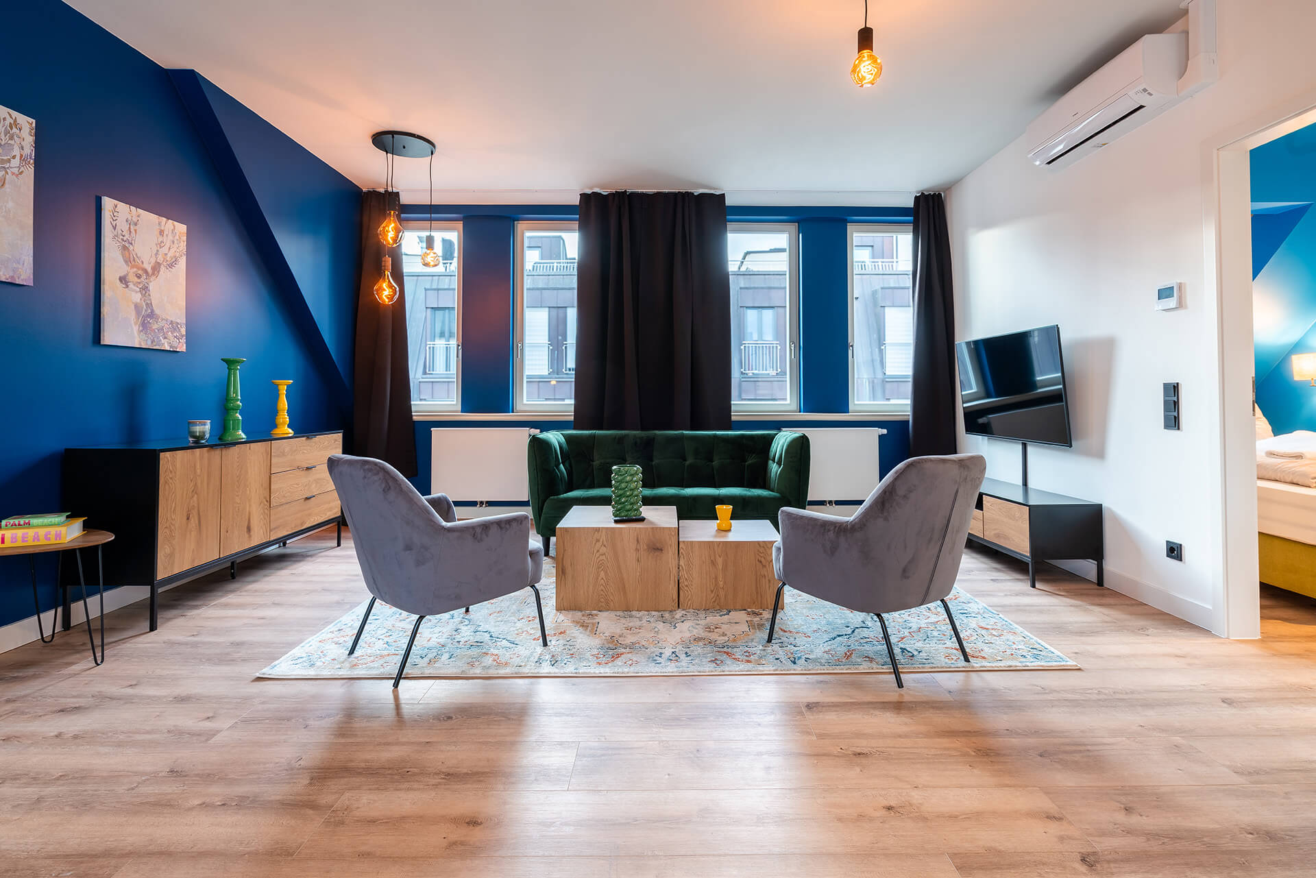 Modern eingerichtetes Apartment namens Lara's Loft im R42 in Leipzig, mit offenem Wohnbereich, grünem Samtsofa, zwei grauen Sesseln, großem Teppich, Wand-TV, hellem Holzfußboden sowie angrenzendem Schlafzimmer.