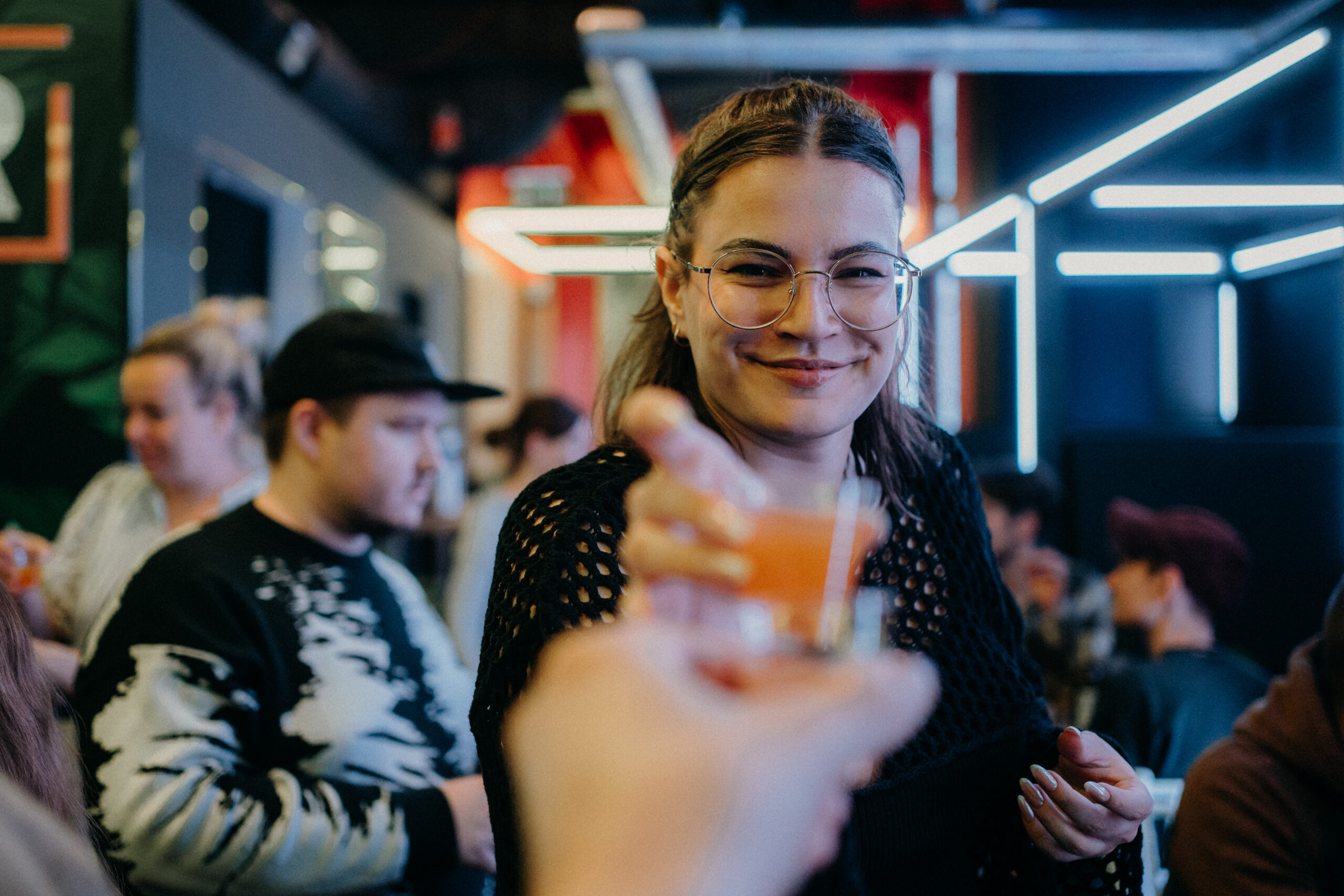 Eine junge Frau guckt frontal Richtung Kamera und erhält von einer anderen Person ein kleines Glas mit einer orangefarbenen Flüssigkeit. Von der anderen Person ist nur die Hand zu sehen, so als würde sie selbst die Kamera halten. Im Hintergrund stehen und sitzen mehrere Personen. Es ist etwas Deko wie Leuchtstoffröhren zu sehen.