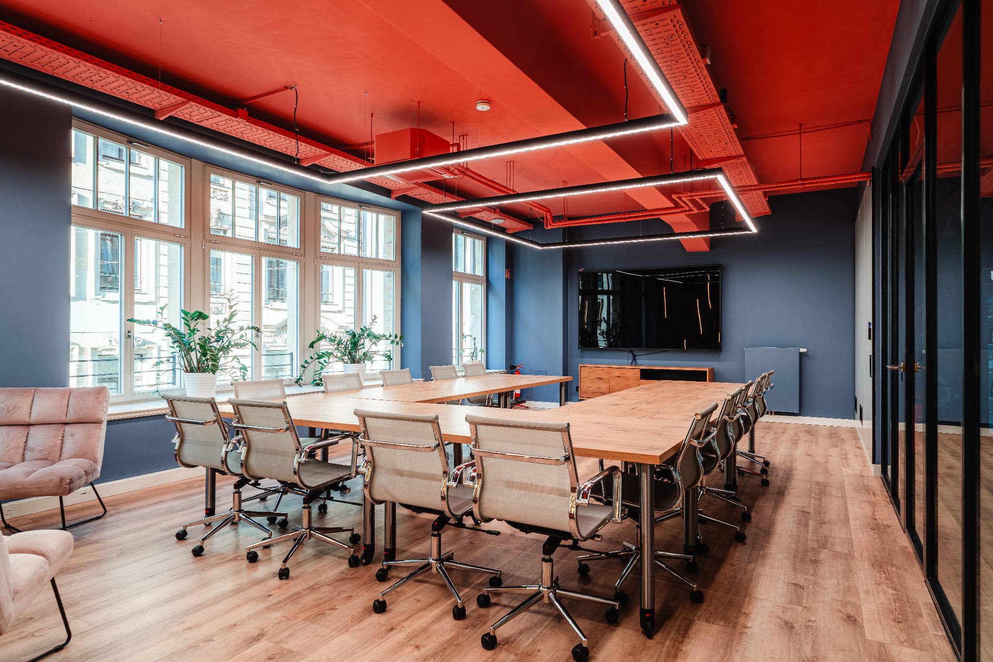 Workshopraum ‚Weitblick‘ im R42 Leipzig: heller, moderner Raum mit großen Fenstern, Holzfußboden, langen Konferenztischen mit Drehstühlen in U-Form gestellt, blauen Wänden, rotem Deckenakzent und linearer LED-Beleuchtung – ideal für Workshops und buchbar.