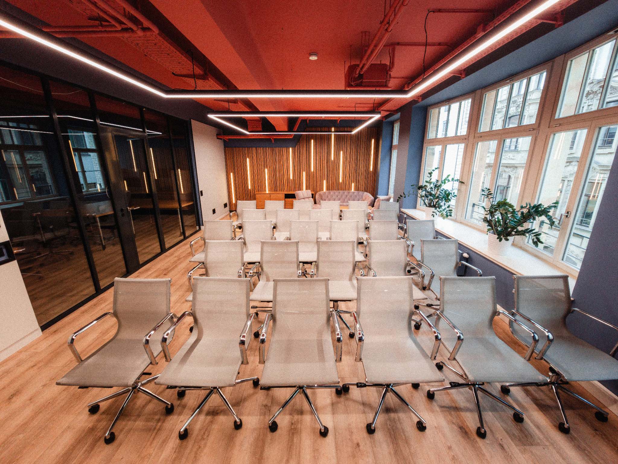 Workshopraum ‚Weitblick‘ im R42 Leipzig: heller, moderner Raum mit großen Fenstern, Holzfußboden, Drehstühlen in Reihen aufgestellt, blauen Wänden, rotem Deckenakzent und linearer LED-Beleuchtung – ideal für Workshops und buchbar.
