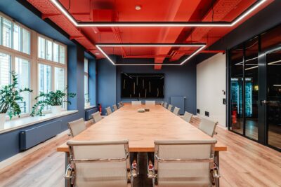 Workshopraum ‚Weitblick‘ im R42 Leipzig: heller, moderner Raum mit großen Fenstern, Holzfußboden, langen Konferenztischen mit Drehstühlen in Block-Form gestellt, blauen Wänden, rotem Deckenakzent und linearer LED-Beleuchtung – ideal für Workshops und buchbar.