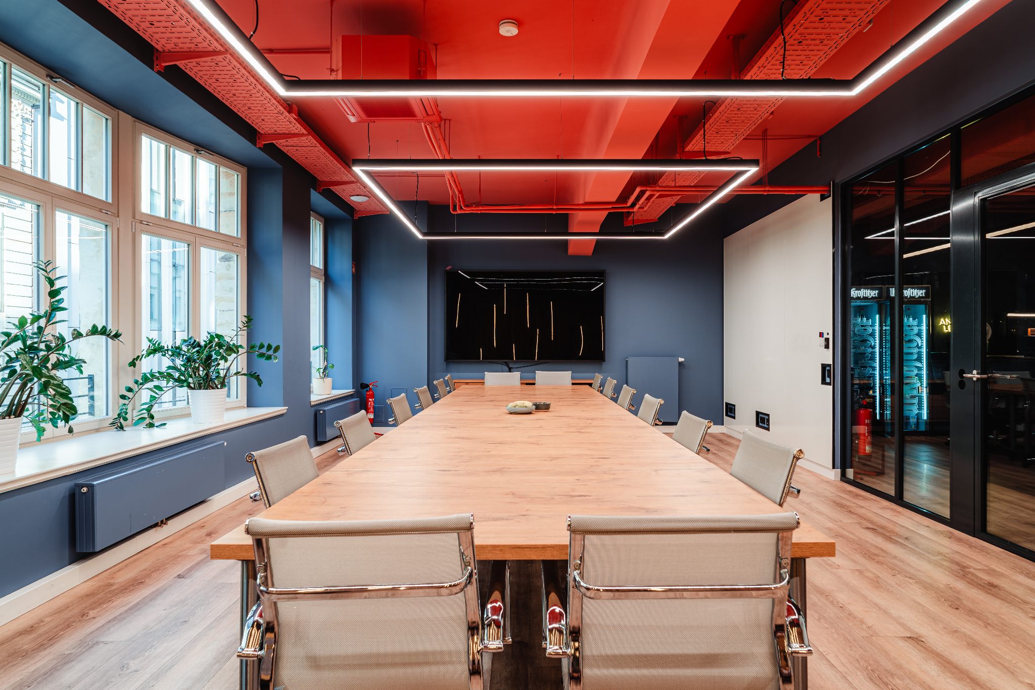 Workshopraum ‚Weitblick‘ im R42 Leipzig: heller, moderner Raum mit großen Fenstern, Holzfußboden, langen Konferenztischen mit Drehstühlen in Block-Form gestellt, blauen Wänden, rotem Deckenakzent und linearer LED-Beleuchtung – ideal für Workshops und buchbar.