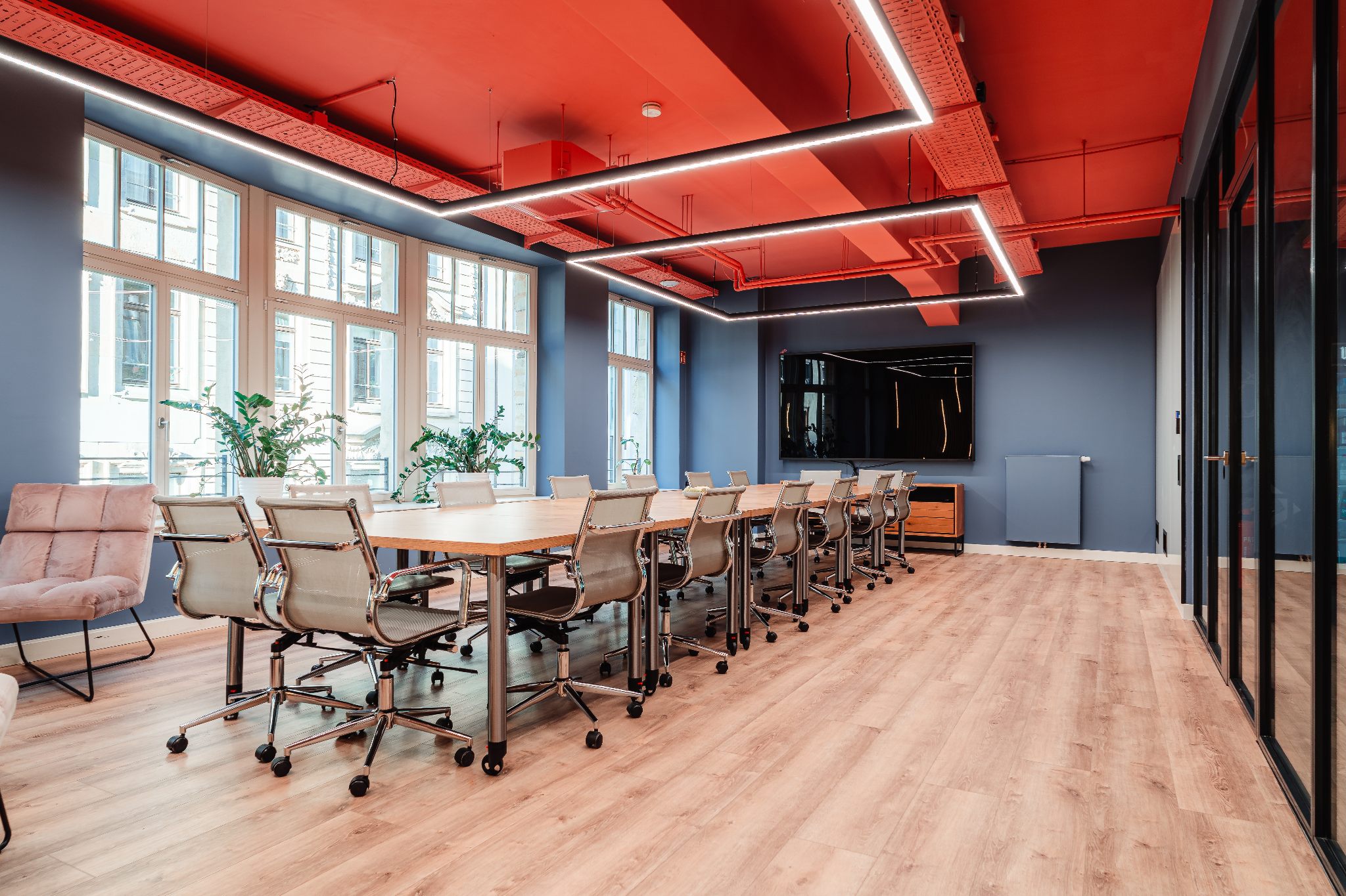 Workshopraum ‚Weitblick‘ im R42 Leipzig: heller, moderner Raum mit großen Fenstern, Holzfußboden, langen Konferenztischen mit Drehstühlen in Block-Form gestellt, blauen Wänden, rotem Deckenakzent und linearer LED-Beleuchtung – ideal für Workshops und buchbar.