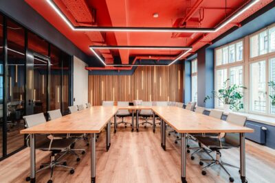 Workshopraum ‚Weitblick‘ im R42 Leipzig: heller, moderner Raum mit großen Fenstern, Holzfußboden, langen Konferenztischen mit Drehstühlen in U-Form gestellt, blauen Wänden, rotem Deckenakzent und linearer LED-Beleuchtung – ideal für Workshops und buchbar.