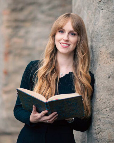 Anabelle Stehl lehnt an einer Steinmauer. Sie trägt schwarze Kleidung, lange offene blonde Haare und hält ein Buch aufgeschlagen in der Hand.
