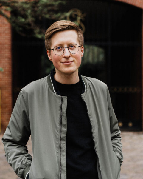 Mikkel Robrahn steht im Freien mit Blick zur Kamera. Er hat kurze braune Haare, trägt ein schwarzes Shirt und eine grau Jacke. Er hat beide Hände in seinen Jackentaschen.