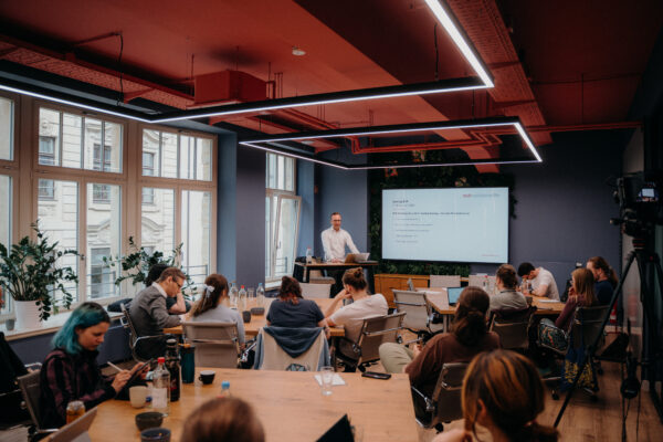 Ein Workshop-Raum im R42 Games Hub mit konzentriert arbeitenden Menschen an mehreren Tischen. Ein Mann in weißem Hemd steht ganz vorn an einem Hochtisch und spricht. Hinter ihm ist eine Präsentation zu Rechtsthemen auf einem großen Bildschirm.